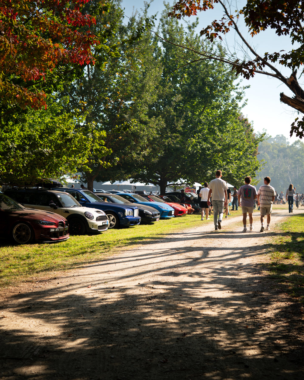 People walking along a dirt path next to parked cars under trees with autumn foliage.